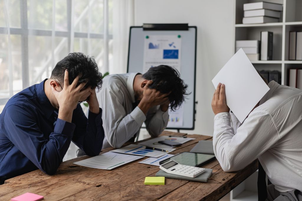 Drei Geschäftsleute sitzen mit verzweifeltem Blick am Tisch und halten ihre Köpfe in den Händen.