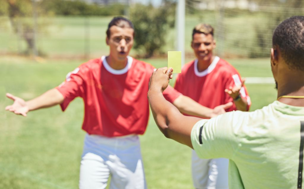 Ein Fussballschiedsrichter hält eine gelbe Karten hoch, um eine Strafe im Spiel zu signalisieren. Zwei Spieler protestieren gestikulierend gegen die Verwarnung.