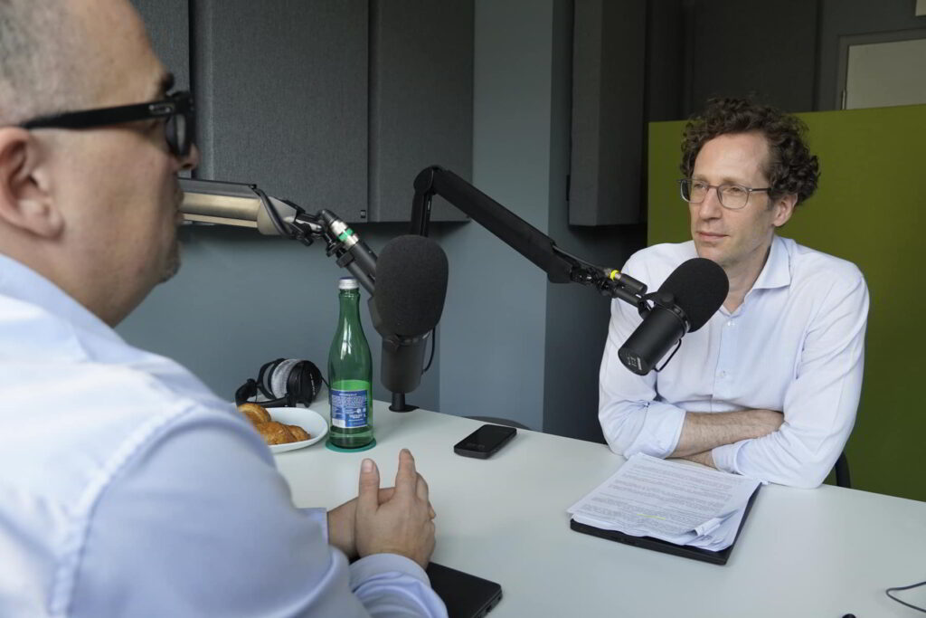 Zwei Personen in einem Tonstudio führen ein Podcast-Interview mit Mikrofonen, Notizen und Kopfhörern auf dem Tisch.