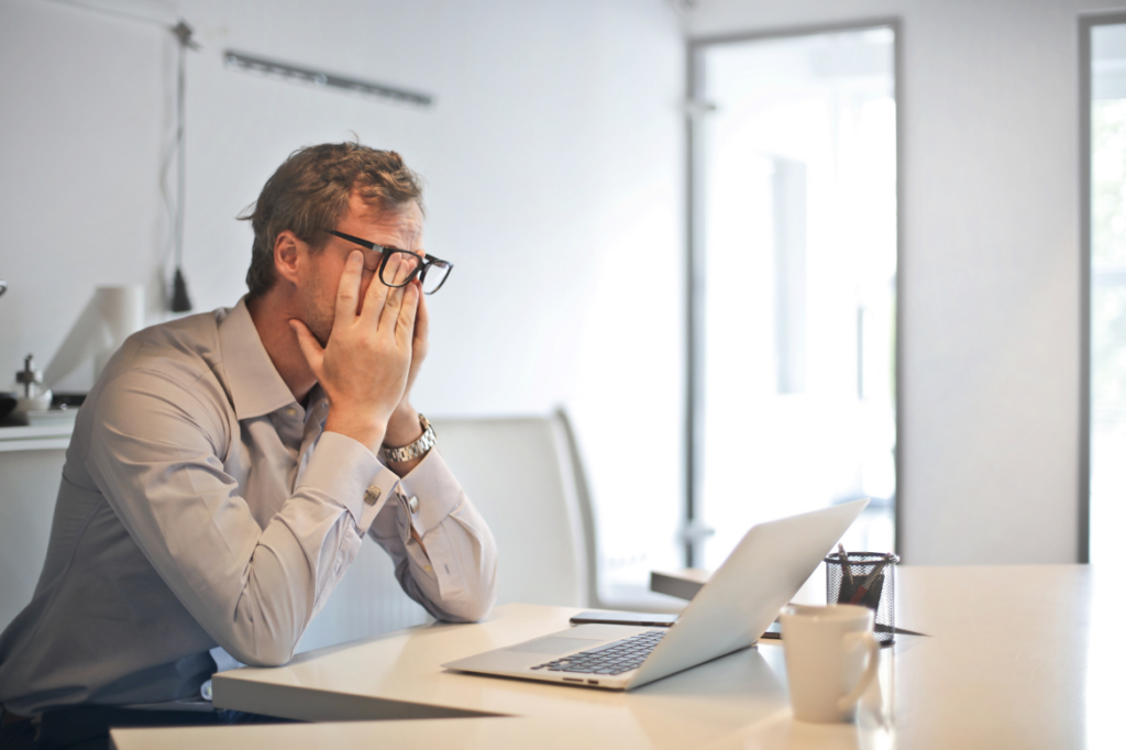 Gestresster Mann im Büro sitzt vor dem Laptop und reibt sich das Gesicht – Ausdruck von Überforderung.