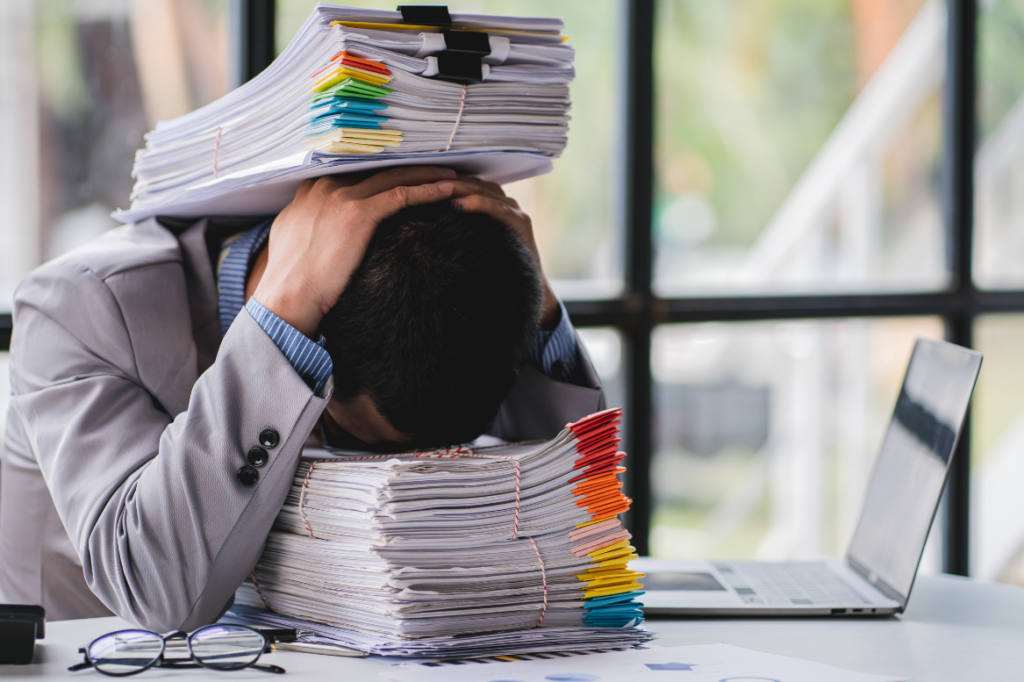 Gestresster Büroangestellter mit dem Kopf auf einem Stapel Aktenordner, zusätzlich belastet durch weitere Akten auf dem Kopf; Symbolbild für Überforderung und Papierflut im Büroalltag.