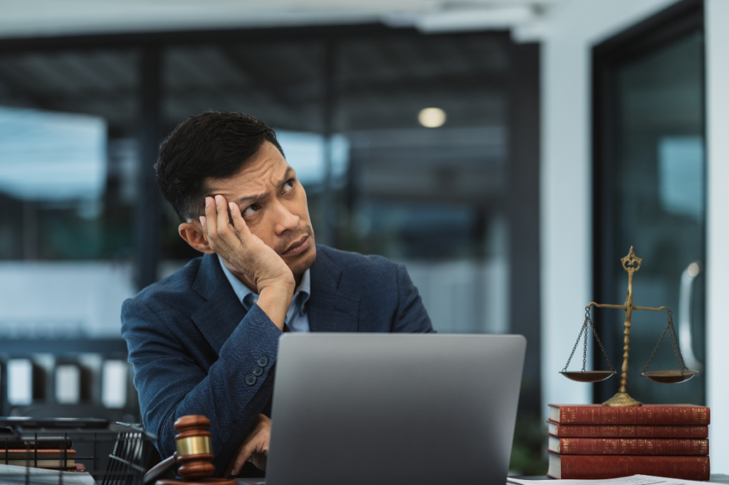 Nachdenklicher Mann in Anwaltskanzlei sitzt vor Laptop mit Justizsymbolen – Ausdruck von Unsicherheit oder Frustration.