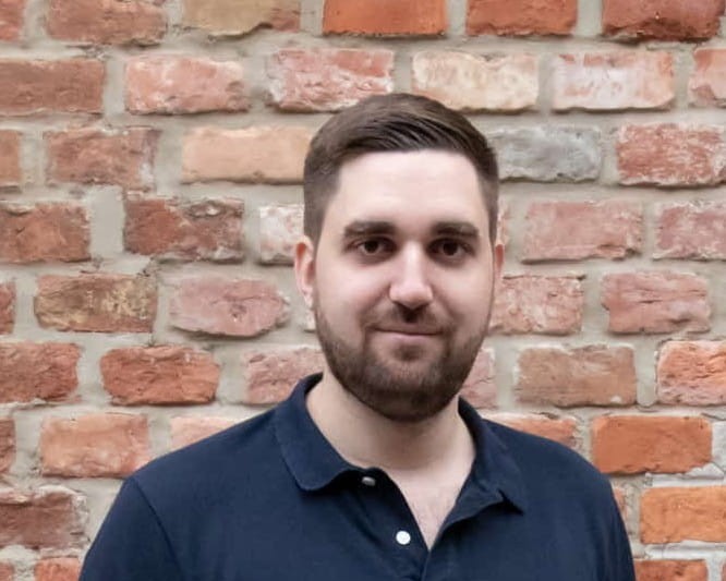 Porträt von Philipp Kamper vor einer Ziegelwand, in dunklem Poloshirt, freundlich in die Kamera blickend.